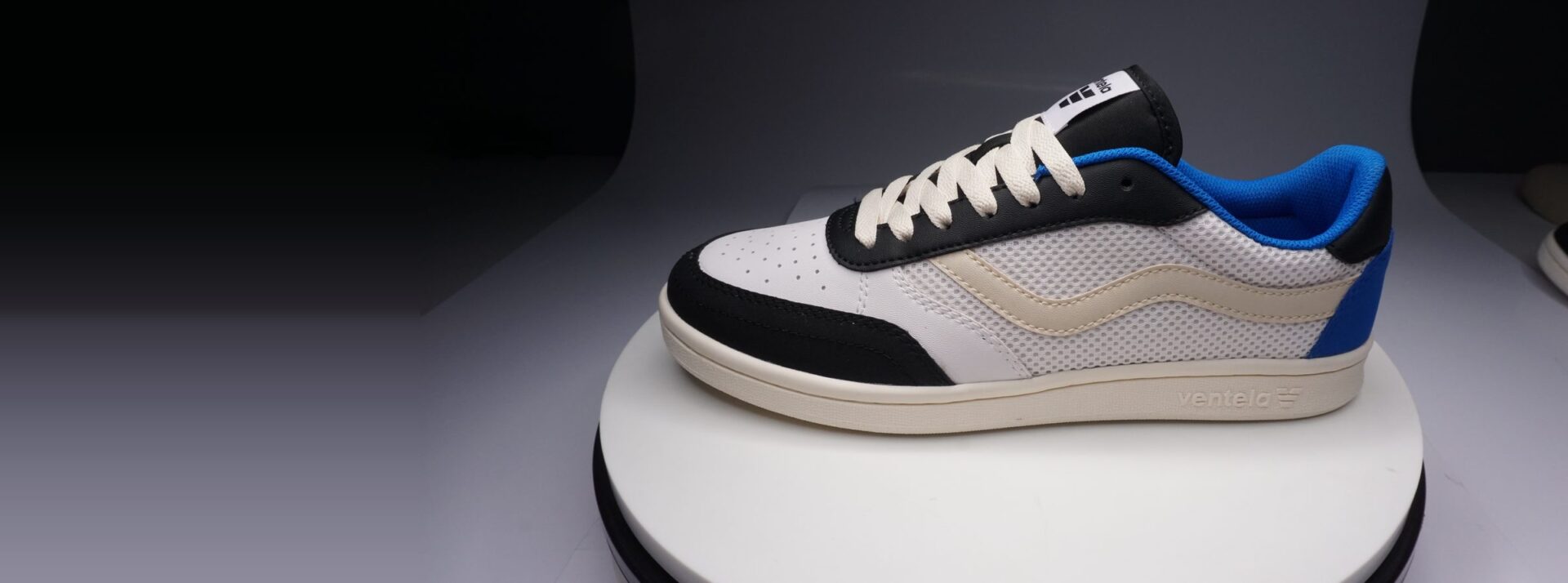 Sepatu Ventela V-88 putih, hitam, dan biru di meja putar studio berlatar gelap